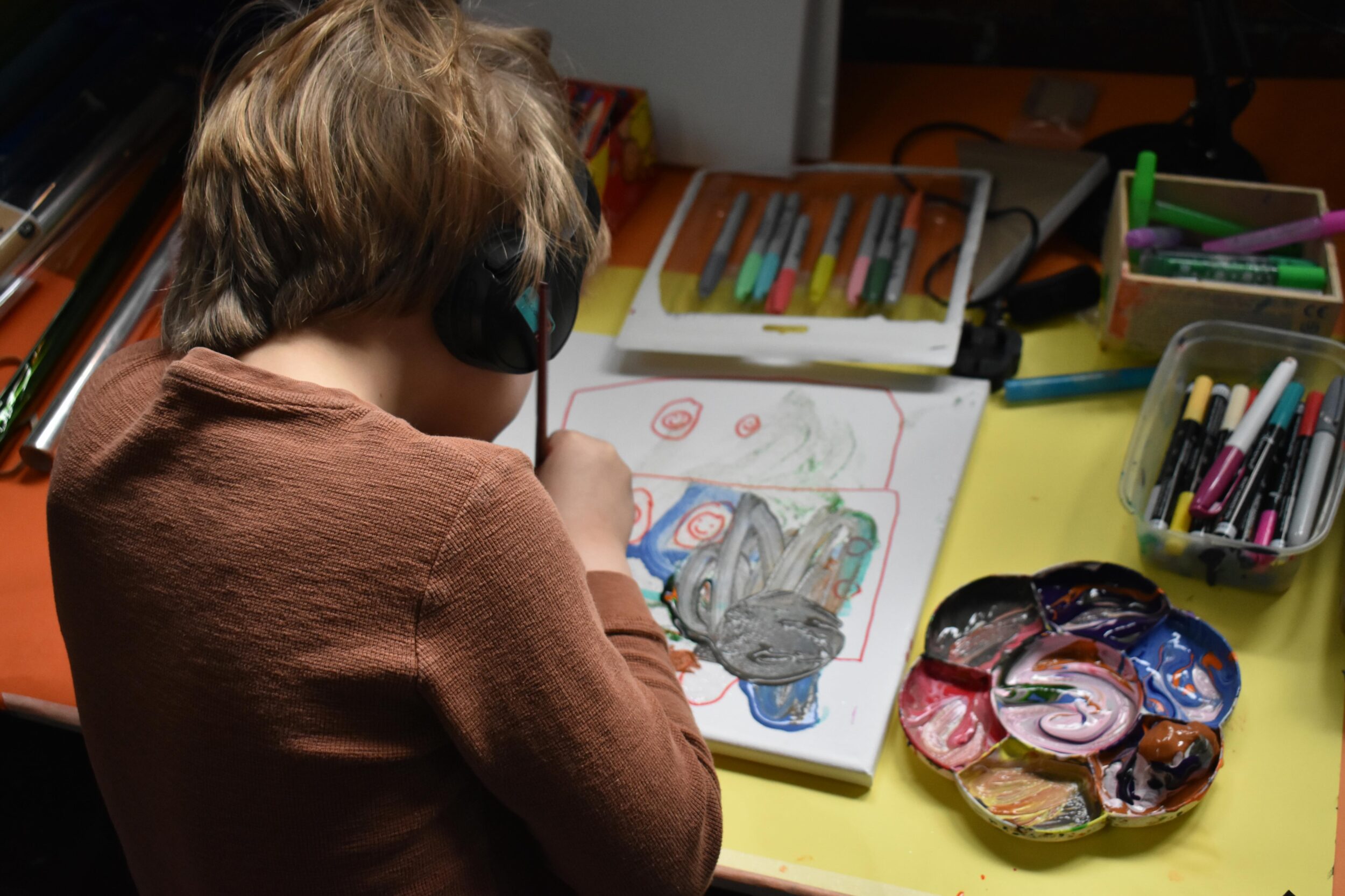 A young child works on a painting. They re surrounded by art supplies. The child is wearing ear defenders.