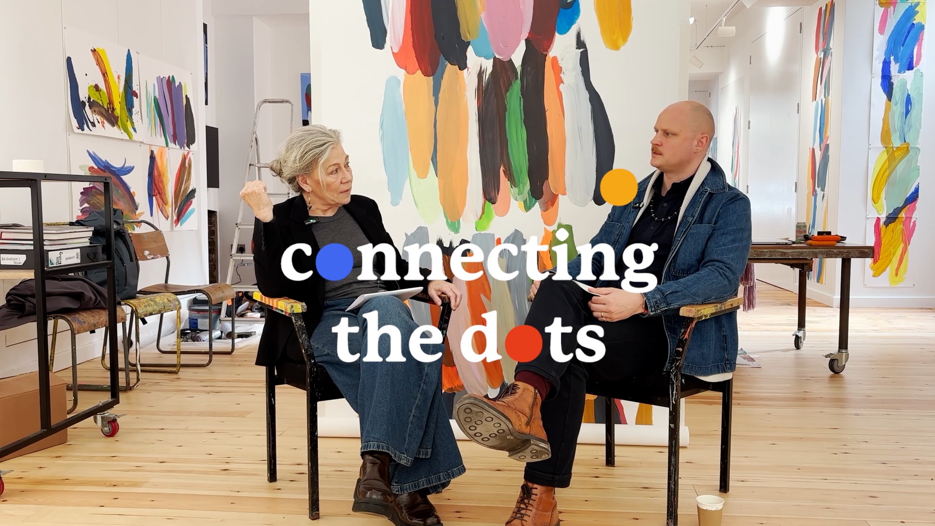 Kate and George sit in the Project art Works gallery at 12 Claremont surrounded by bright, beautiful paintings by Christopher Tite. They sit side by side chatting.